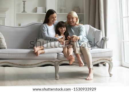 Full length happy 3 generations family sitting together on couch in living room, watching funny cartoons, recording video, taking selfie. Smiling little girl enjoying free time with mommy and grandma.