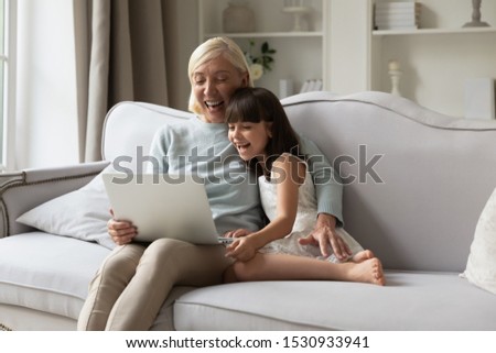 Overjoyed middle aged woman embracing small preschool happy granddaughter, watching funny comedian movie on laptop, laughing, enjoying leisure weekend time together sitting on cozy sofa at home.