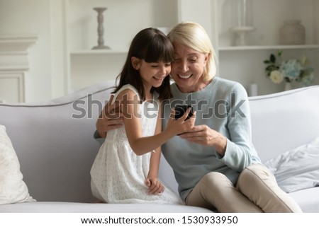 Overjoyed mature grandmother cuddling little granddaughter, watching funny video together on smartphone, sitting on comfortable couch at home. Happy girl with granny holding video call on cellphone.