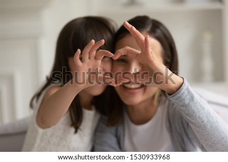 Happy young mother with cute little daughter making focused heart sign with hands, looking at camera. Smiling millennial mom and small girl showing love gesture together, expressing care, affection.