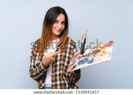 Young painter woman over isolated blue background and pointing it