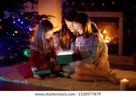 Happy young mother and her two small daughters opening a magical Christmas gift by a fireplace in a cozy dark living room on Christmas eve. Winter evening at home with family and kids.