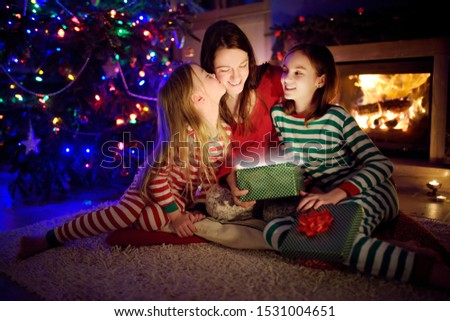 Happy young mother and her two small daughters opening a magical Christmas gift by a fireplace in a cozy dark living room on Christmas eve. Winter evening at home with family and kids.