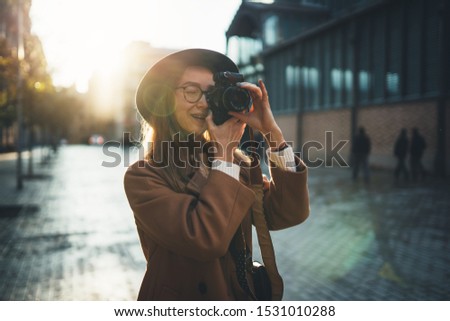 Outdoor smiling lifestyle portrait of pretty young woman having fun in sun city in Europe with camera travel photo of photographer Making pictures in hipster style glasses and hat 