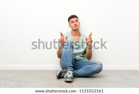 Young handsome man sitting on the floor with fingers crossing