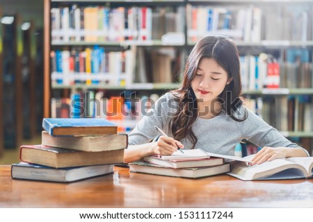 Asian young Student in casual suit reading and doing homework in library of university or colleage with various book and stationary on the wooden table over the book shelf background, Back to school Royalty-Free Stock Photo #1531117244