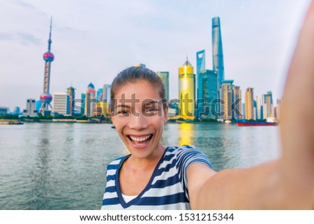 Selfie Asian tourist chinese woman China travel. Smiling young girl excited holding smartphone camera to take a picture with phone of herself in front of Shanghai's skyline of skycrapers.