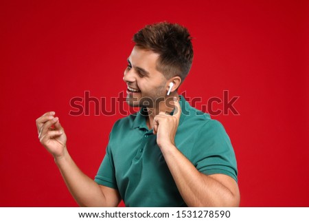 Happy young man listening to music through wireless earphones on red background