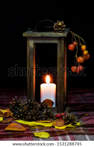 Composition of a burning candle in an old lantern with fallen autumn leaves, pine cones and apples on a checkered plaid.