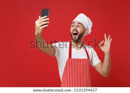 Excited young chef cook or baker man in striped apron toque chefs hat isolated on red wall background. Cooking food concept. Mock up copy space. Doing selfie shot on mobile phone, showing OK gesture