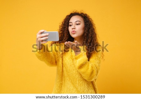 Pretty young african american girl in fur sweater posing isolated on yellow orange background. People lifestyle concept. Mock up copy space. Doing selfie shot on mobile phone blowing sending air kiss