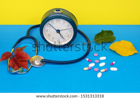 The concept of autumn colds. Alarm clock for the timely adoption of pills, a stethoscope, multi-colored pills, autumn leaves of different colors on a blue background.