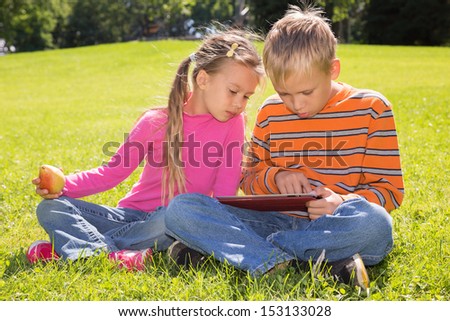 A boy and a girl are looking at the screen of a tablet computer while lying on a green lawn