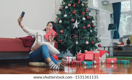 Asian women celebrate Christmas festival. Female teen relax happy holding Gift and using smartphone selfie with Christmas tree enjoy xmas winter holidays in living room at home.