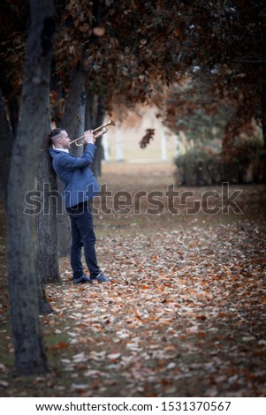 slender attractive  male musician in blue jacket stands in autumn park alley of fall trees of brown and yellow colours and plays golden old trumpet 