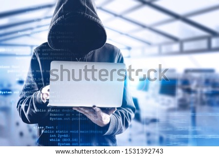 Young unrecognizable hacker in black hoodie using laptop over blurry office background with double exposure of lines of code. Concept of identity theft and cyber security. Toned image