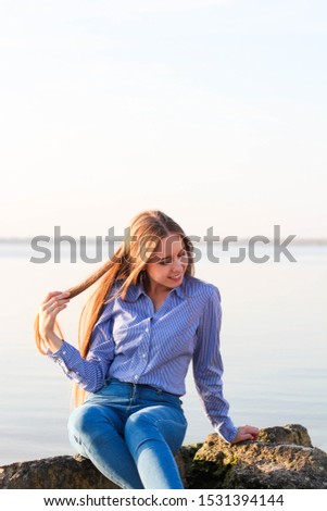 Beautiful smiling girl with long hair in shirt and jeans sits on stones on beach on sunny day.
