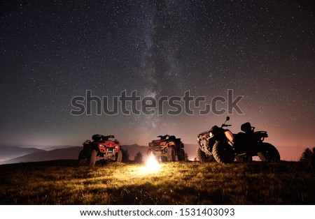 Night picture. Three atv quad motorbikes standing on the top of mountain near burning bonpfire, under amazing night starry sky and Milky way on background