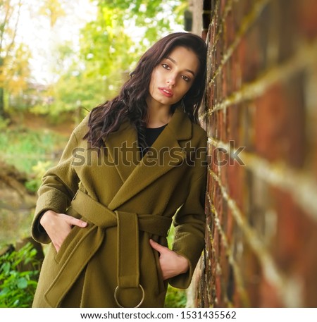 photo of the girl at the wall. Young woman wrapped in a khaki coat.portrait of a girl with long gorgeous brunette hair in a green coat  or against a vintage red brick wall that goes to perspective