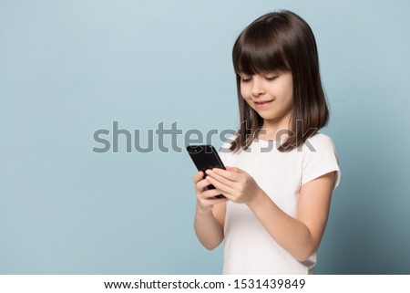 Cute preschooler little girl stand isolated on blue studio background using modern smartphone, smart small kid browsing surfing internet watch cartoon on cellphone, easy access, blank copy space