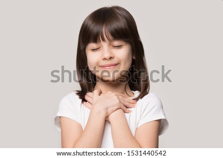 Close up of cute happy small girl isolated on grey studio background hold hands at heart chest feel grateful, smiling little child with eyes closed pray thanking god high powers, faith concept