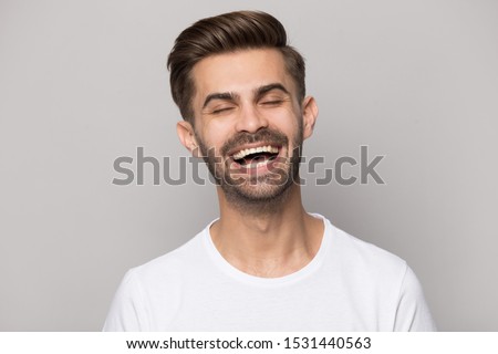 Headshot portrait of happy Caucasian bearded millennial guy isolated on grey studio background laughing at funny joke, smiling overjoyed man in white t-shirt have fun, entertainment, humor concept
