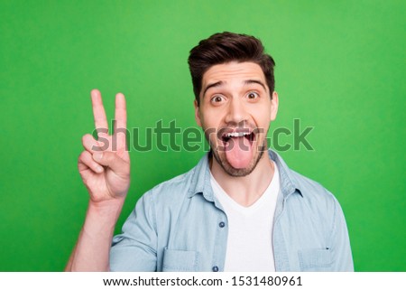 Closeup photo of amazing guy showing v-sign symbol sticking tongue out mouth wear casual denim shirt isolated over green color background