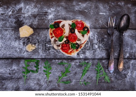 Preparation of heart-shaped pizza from ingredients such as dough, tomatoes, olives, basil, cheese, on a dark wooden board and pressed with flour next to the spoon and fork. Top view.