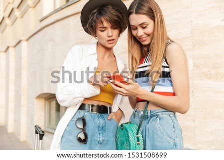 Photo of two concentrated tourist girls looking at smartphone while walking with suitcases on city street