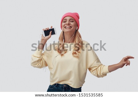 Attractive young woman listening music and smiling while standing against grey background