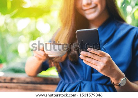 Closeup image of a beautiful asian woman holding and using mobile phone while drinking coffee in the garden