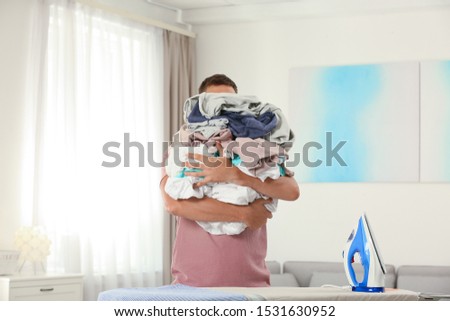 Handsome man with iron and unfolded laundry near board at home