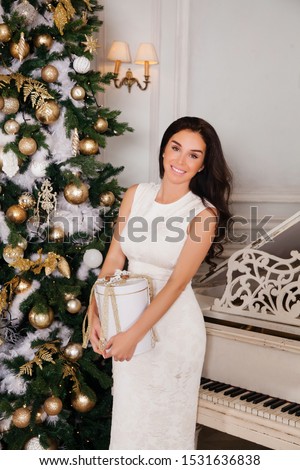 Merry Christmas and Happy Holidays. Beautiful young brunette woman in white dress posing against the background of a white piano and decorated Christmas tree with presents 