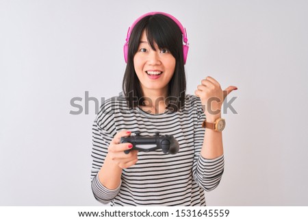 Chinese gamer woman playing video game using headphones over isolated white background pointing and showing with thumb up to the side with happy face smiling