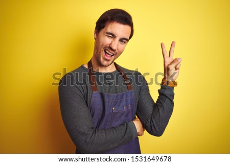 Young handsome shopkeeper man wearing apron standing over isolated yellow background smiling with happy face winking at the camera doing victory sign. Number two.