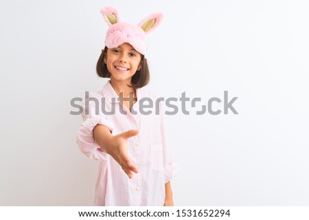 Beautiful child girl wearing sleep mask and pajama standing over isolated white background smiling friendly offering handshake as greeting and welcoming. Successful business.