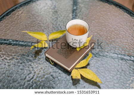 Close up outside photo of book and cup of hot tea with steam on the table. Yellow leaves fall. Warm days, mellow autumn. Sunday relax. Autumnal mood, fall season concept. Selective focus, top view. 