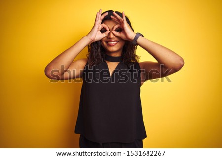 Transsexual transgender elegant businesswoman standing over isolated yellow background doing ok gesture like binoculars sticking tongue out, eyes looking through fingers. Crazy expression.