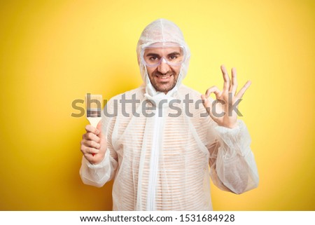 Young man wearing painter equipment and holding painting brush over isolated yellow background doing ok sign with fingers, excellent symbol