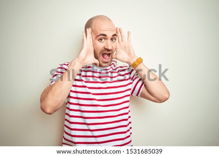 Young bald man with beard wearing casual striped red t-shirt over white isolated background Smiling cheerful playing peek a boo with hands showing face. Surprised and exited