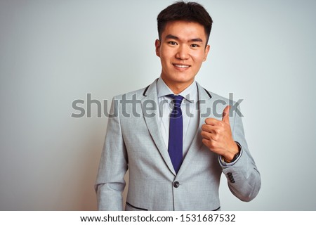 Asian chinese businessman wearing grey jacket and tie standing over isolated white background doing happy thumbs up gesture with hand. Approving expression looking at the camera showing success.