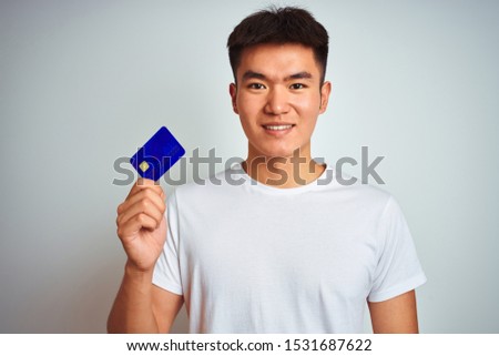 Young asian chinese man holding credit card standing over isolated white background with a happy face standing and smiling with a confident smile showing teeth