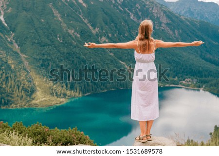 Young blonde in white dress standing at stone in Polish Tatra with Morskie oko lake on background