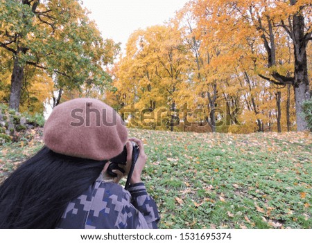  Back of a woman photographer wearing hat taking photograph of a park in autumn with still green lawn with trees full of yellow golden leaves