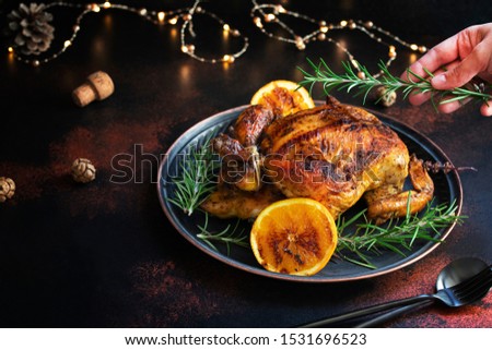 Roasted whole chicken for Christmas or New Year on a dark background with Christmas decorations. Baked Chicken with Oranges. Female hand decorating chicken with rosemary