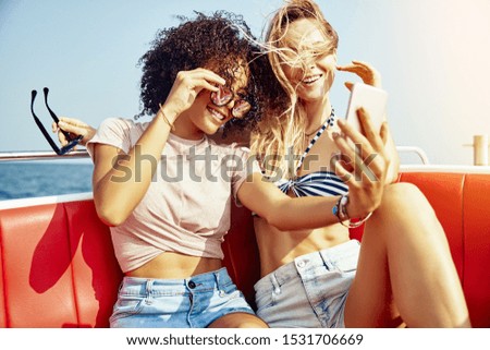 Two smiling young women sitting on a boat on the ocean taking selfies together during their summer vacation