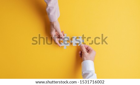 Top view of male and female hands joining two matching puzzle pieces together in a conceptual image. Over yellow background. Royalty-Free Stock Photo #1531716002