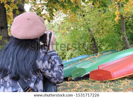Back of a woman photographer wearing hat taking photograph of upside down boats red green and blue on green lawn under trees full of yellow golden leaves  