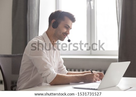 Smiling businessman in headset using laptop, looking at screen, employee student in headphones with microphone watching webinar, making notes, studying online, interpreter translating course