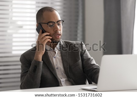 Serious African American businessman in glasses talking on phone, using laptop, having business conversation, hr manager holding interview by cellphone, employee consulting client, making sell offer Royalty-Free Stock Photo #1531731602